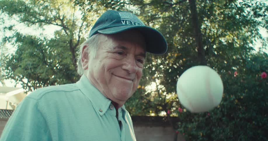 Elderly man standing outdoors with a small american flag throws a baseball into the air and catches it