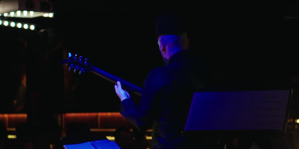 A man in low lighting tunes or prepares his guitar during a stage performance
