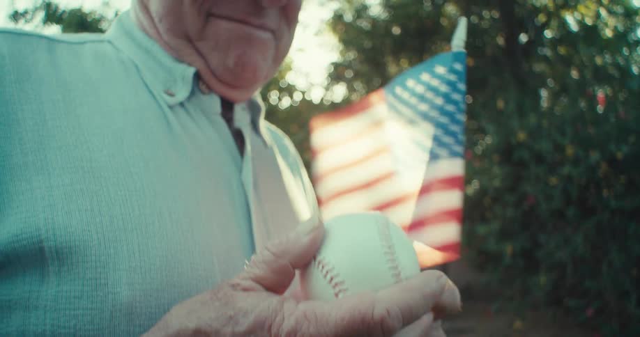 An elderly man gently smiles while holding a baseball near an american flag outdoors