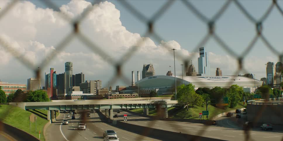 Through a blurred foreground fence a sprawling city skyline featuring a large stadium and modern high rises rises behind a busy highway intersection under a partly cloudy sky