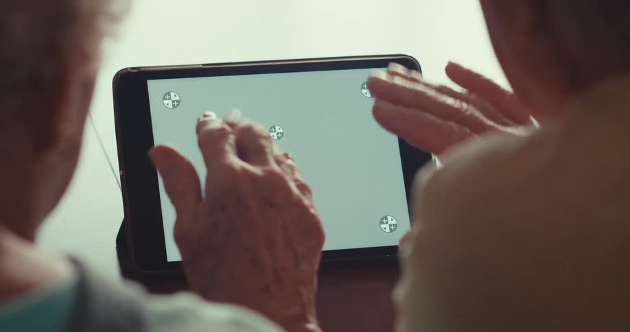 An elderly couple sits side by side indoors waving at a tablet on their desk during a video call