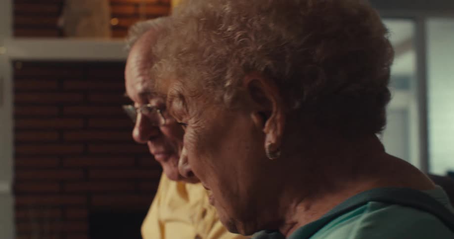 An elderly couple sits indoors while talking and looking at a laptop screen