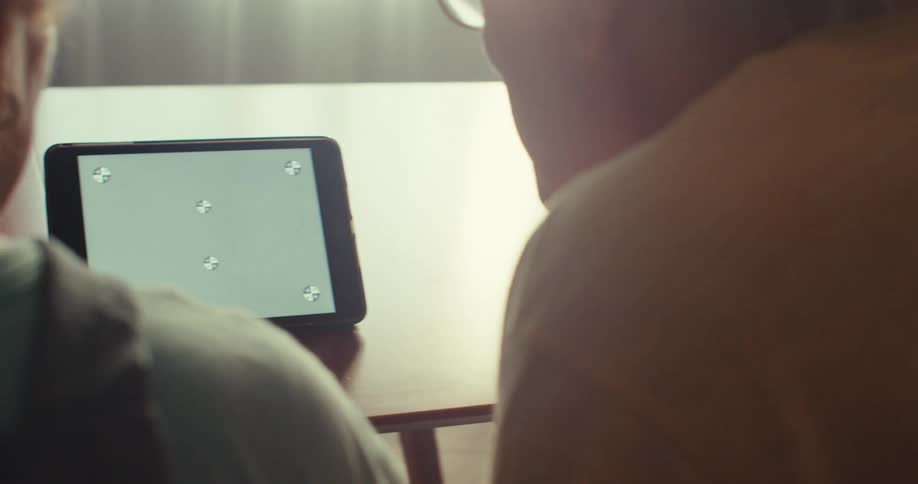 Two people sit closely at a table viewing a greenscreen tablet