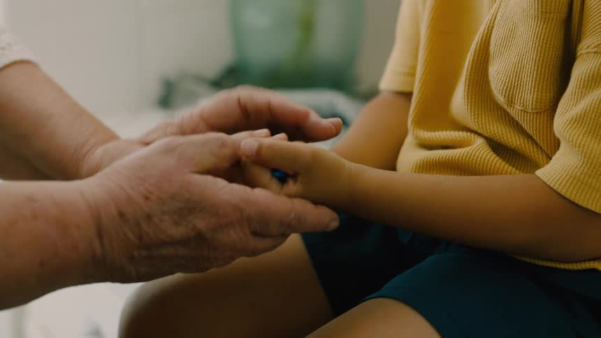 An older hand gently holds a younger hand passing a blue toy into its grasp