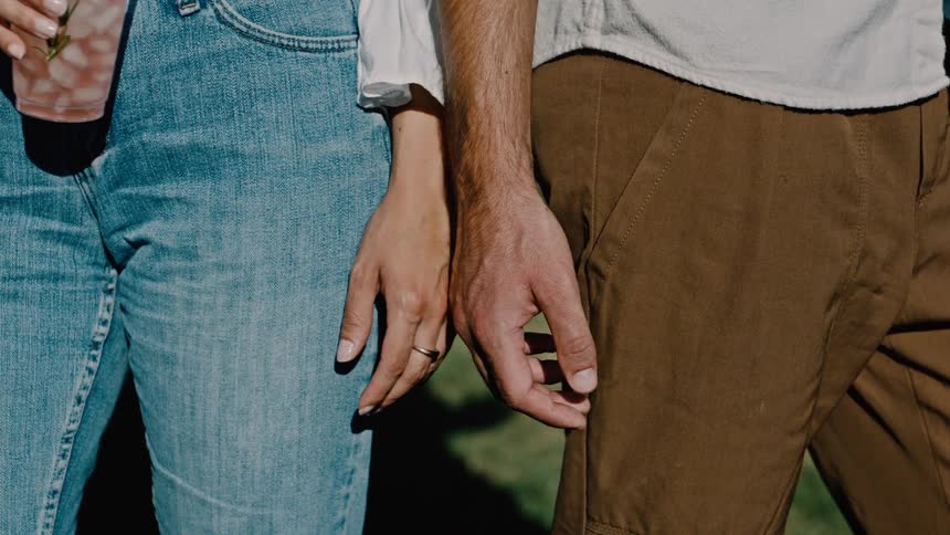Two individuals one in jeans holding a pink drink and the other in brown pants as their hands slowly move towards each other nearly touching