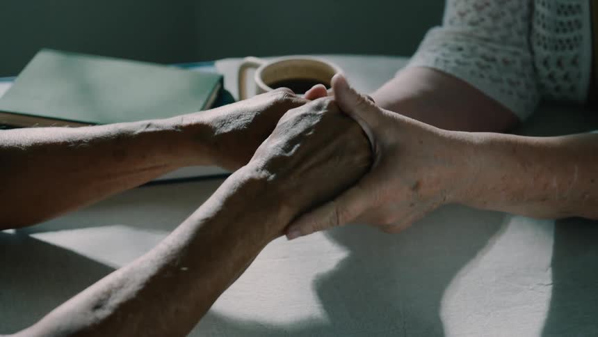 Elderly man and woman hold hands romantically over a tabletop in a room