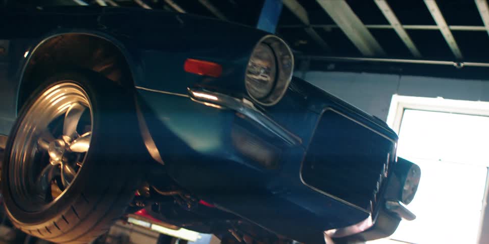 A dark blue vintage car is elevated on a lift in a garage with sunlight streaming in through a window on the right