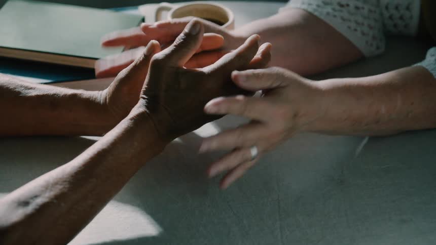 Two people are holding hands on a lightcolored table with a book and a mug in the background