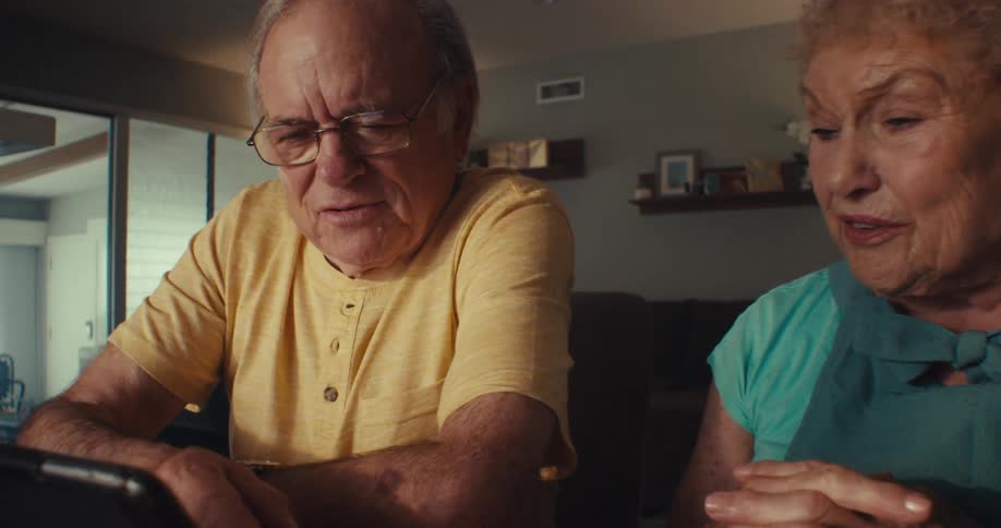 An elderly couple of a man and a woman who are talking and when they are looking at a tablet that is resting on a table