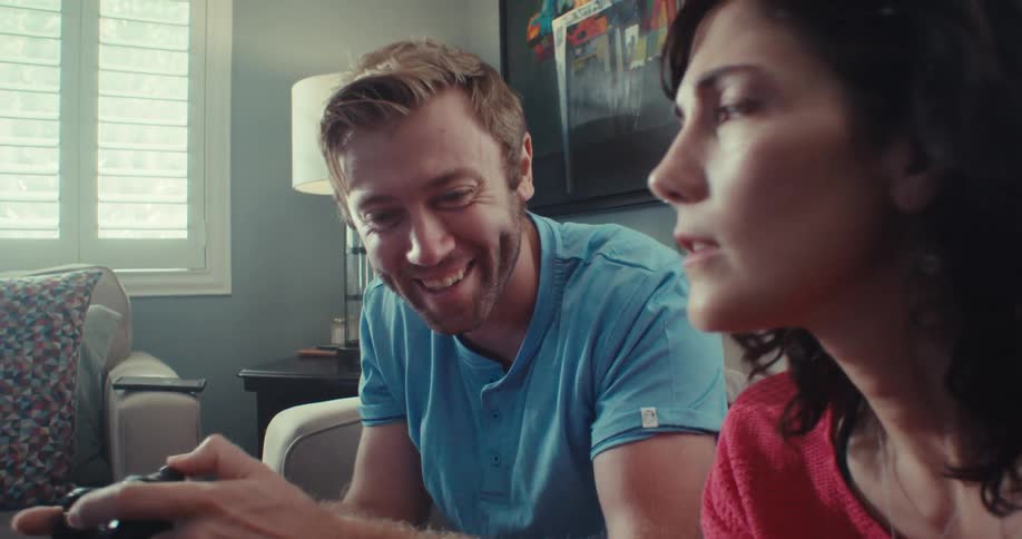 A young couple sits on a couch in a brightly sunlit room engrossed in a video game together
