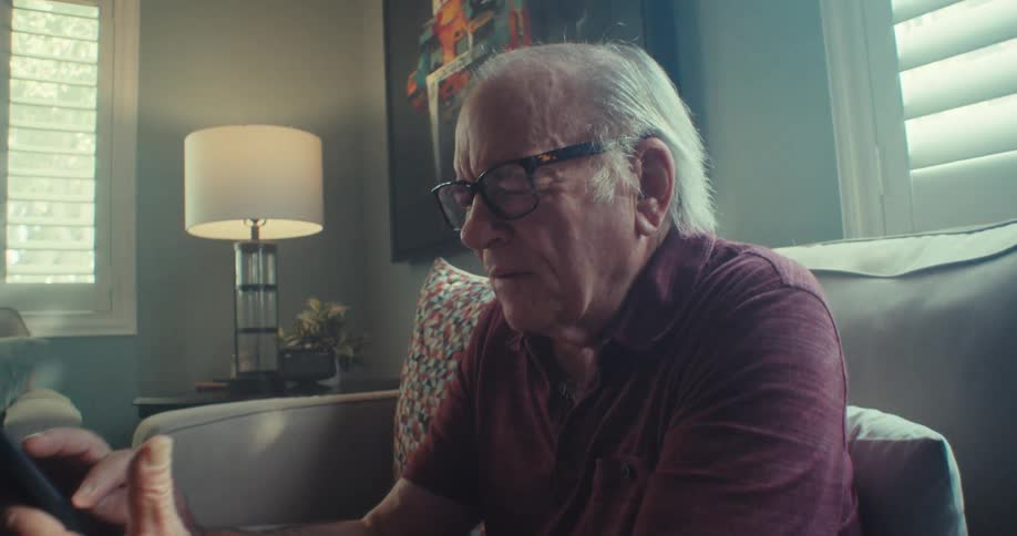 An elderly man sits on a couch holding a smartphone during a video call