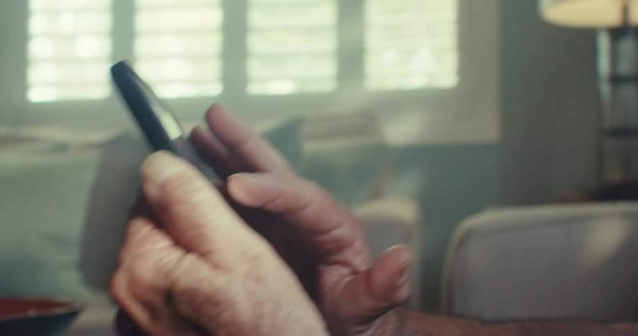 An elderly man wearing glasses sitting on a couch while using a mobile device