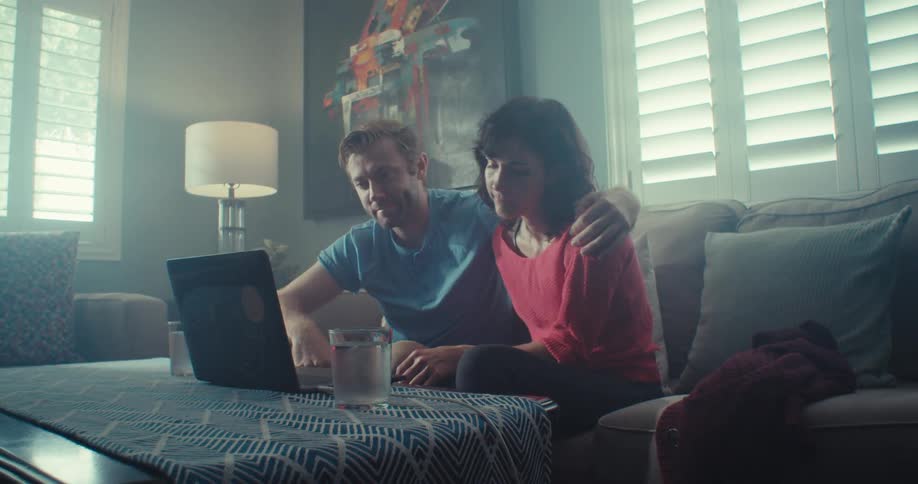 A man has his arm around a womans shoulder as they both look at a laptop together on a couch in a sunlit living room