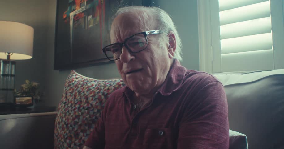An elderly man wearing glasses is sitting on a sofa using a cell phone and making a surprised expression