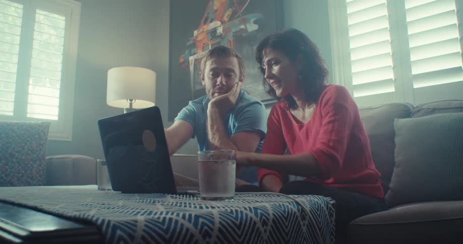 Couple sits on a couch smiling while looking at a laptop and chatting