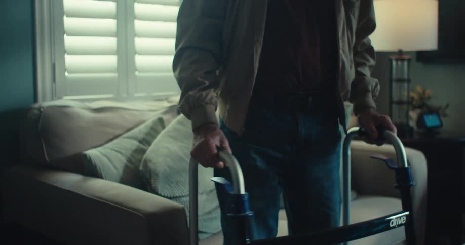 The face of an elderly man who is putting on reading glasses and sitting on a sofa and then he stands up using a walker