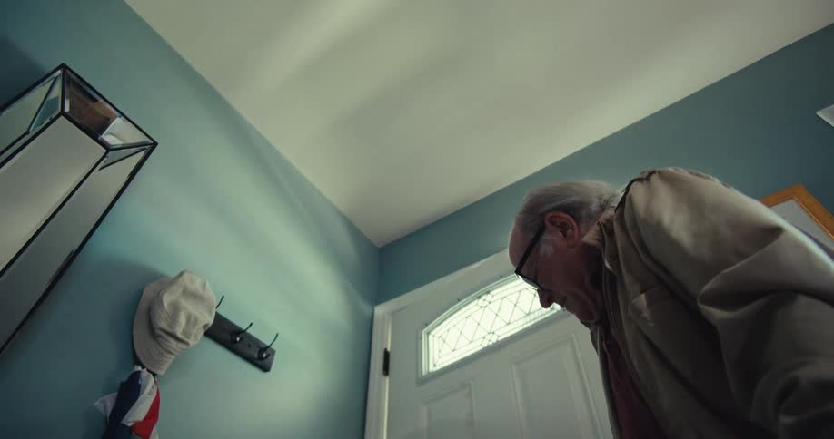 An older man in glasses a lightcolored jacket and a reddish shirt enters a home closing the door behind him on a bright day viewed from the perspective of his walker