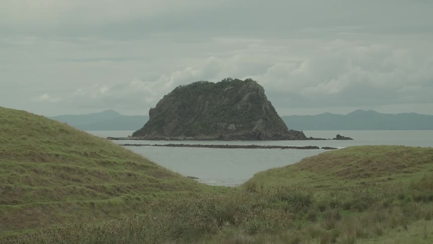 A small rocky island rises from a calm sea framed by grassy hills under an overcast sky