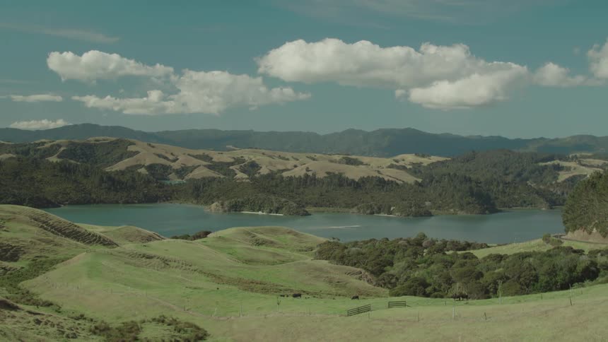 Rolling green hills overlook a peaceful lake under a bright blue sky with scattered clouds