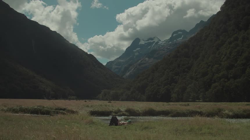 A woman lying on the grass in a wide grassland near a stream surrounded by a vast range of mountains under the bright midday sun