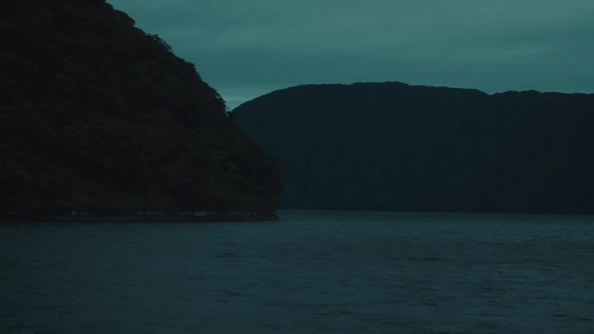 A calm body of water is surrounded by dark treecovered mountains under a murky overcast sky