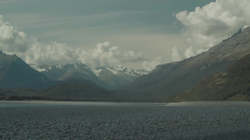A lake sits below snowy mountains under dramatic clouds