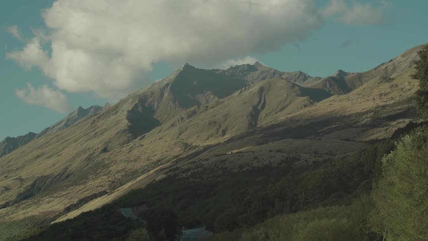A rugged mountain range under a partly cloudy sky with a winding road visible at the base
