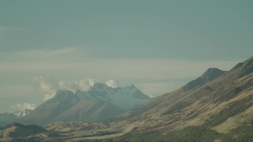 A mountain range with snowcapped peaks in the distance is viewed across a wide valley under a hazy partly cloudy sky