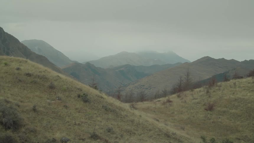 Dry grassy rolling hills lead into a valley with hazy distant mountains under an overcast sky