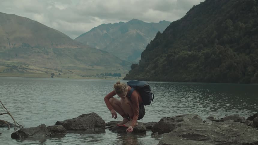 Woman crouches by lake touching water with mountains under cloudy sky