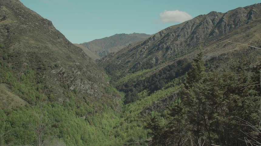 A sunlit mountain valley densely covered with trees is flanked by steep slopes under a partly cloudy sky