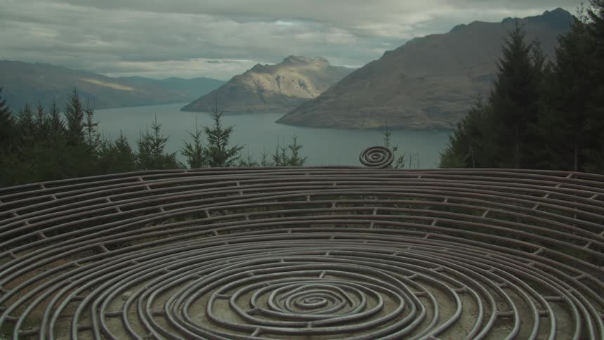 A spiralshaped metal sculpture overlooks a scenic lake surrounded by mountains and dense forest
