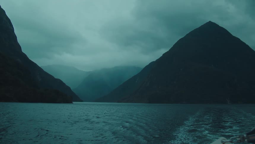 A boat travels through a dark narrow fjord surrounded by steep mountains under a cloudy sky