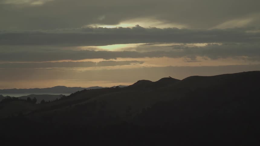 Golden sunset light breaks through dramatic clouds over silhouetted rolling hills
