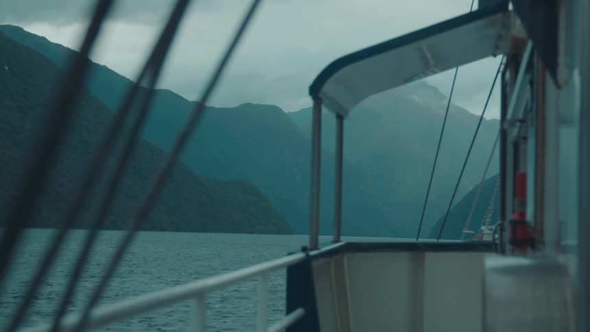 A woman in a dark jacket walks along the deck of a boat offering a view of a majestic fjord and surrounding mountains under an overcast sky