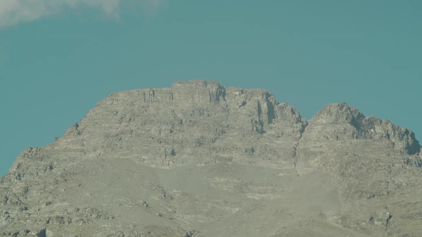 A rugged barren mountain peak with visible rock strata stands sharply against a clear blue sky with a small wisp of cloud