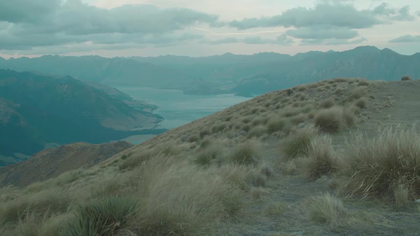 Wind sweeps across a grassy mountainside overlooking a vast valley with a distant lake and mountain ranges under a cloudy sky