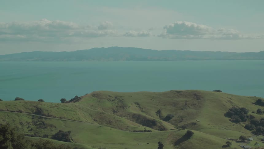 A panoramic view shows green rolling hills in the foreground with a wide body of water stretching to distant mountains under a partly cloudy sky