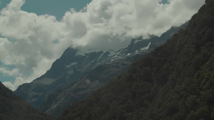 A majestic cloudkissed mountain partially covered in snow towers over a dark treefilled valley under a bright sky