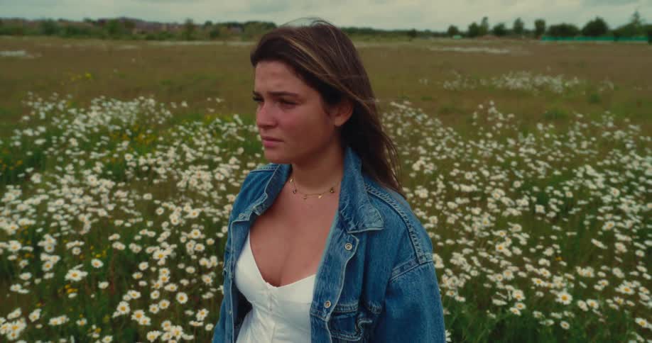 A young woman stands in a field of white daisies under a cloudy sky as the wind lifts her hair