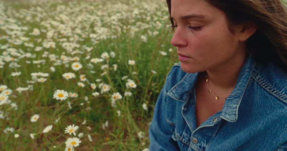 A teenage girl standing in a flowerfilled field at midday wearing a jacket and looking sad as the wind gently blows her hair