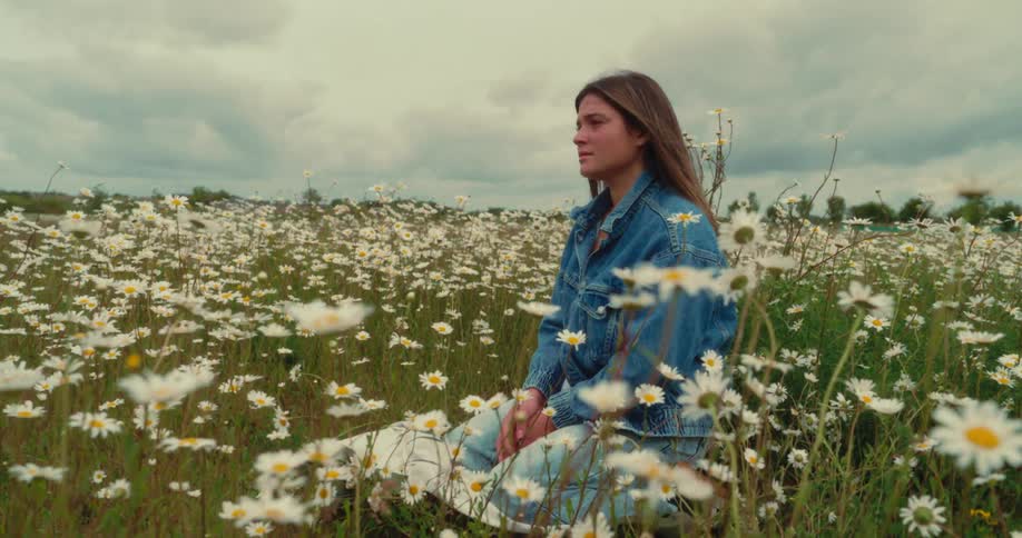 A young woman sits calmly in a blooming daisy field under a cloudy sky looking thoughtful