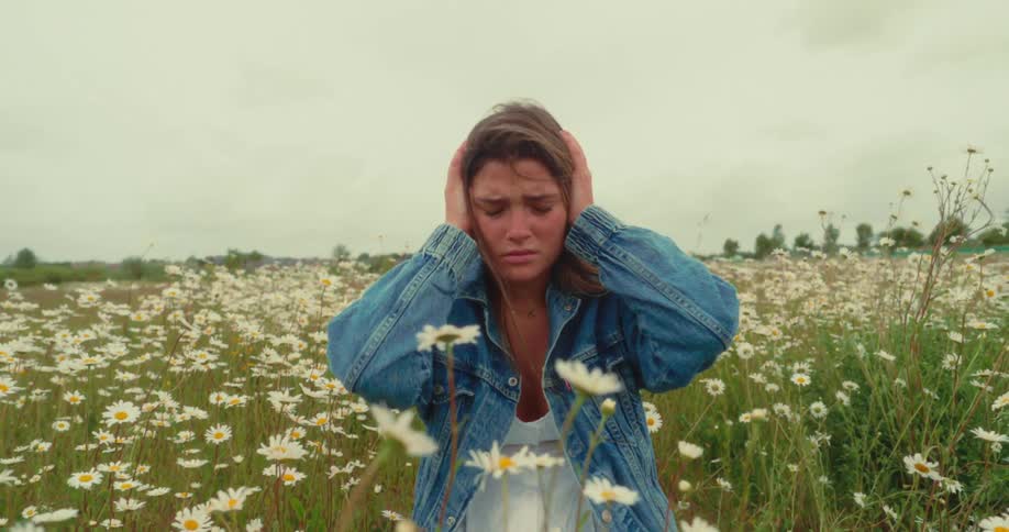 Young woman covers her ears contemplatively while kneeling among wildflowers in a meadow during the day