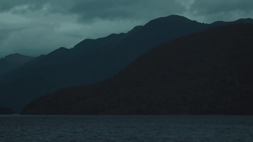 Dark silhouetted mountains rise steeply from a body of water with a moody overcast sky above them