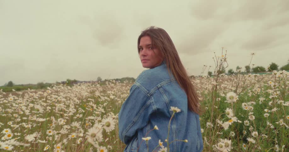 A young woman wearing a denim jacket is looking over her shoulder in a field filled with white daisies under a cloudy sky