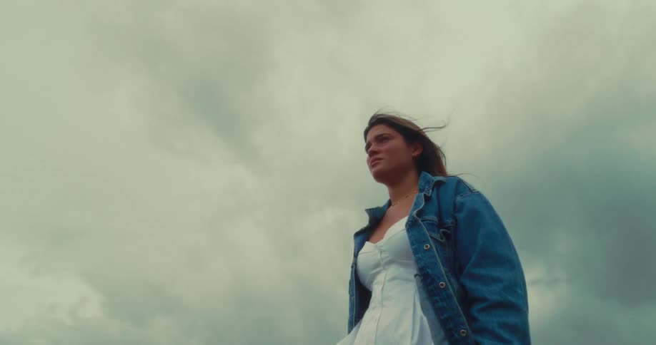 A woman stands staring straight ahead in an outdoor setting with a cloudy sky