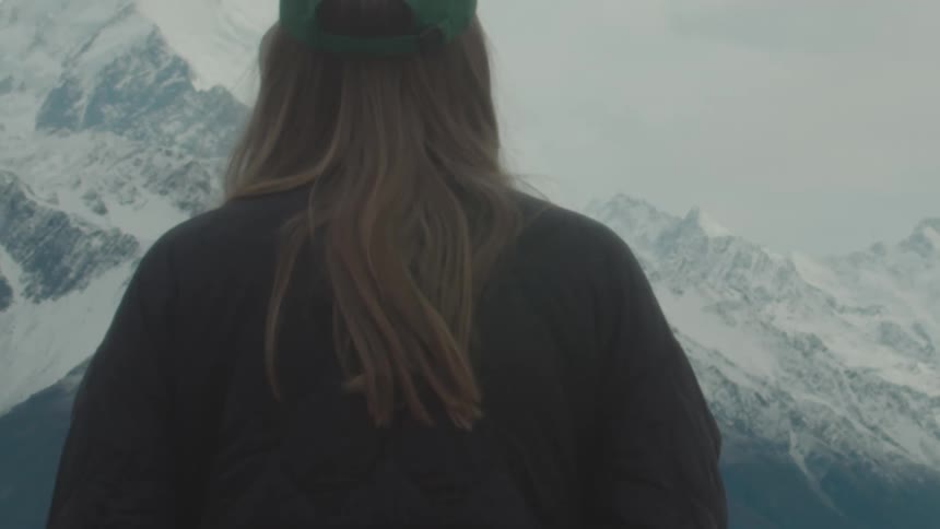 A woman with blonde hair stands with her back facing a mountain range with snowcapped mountains