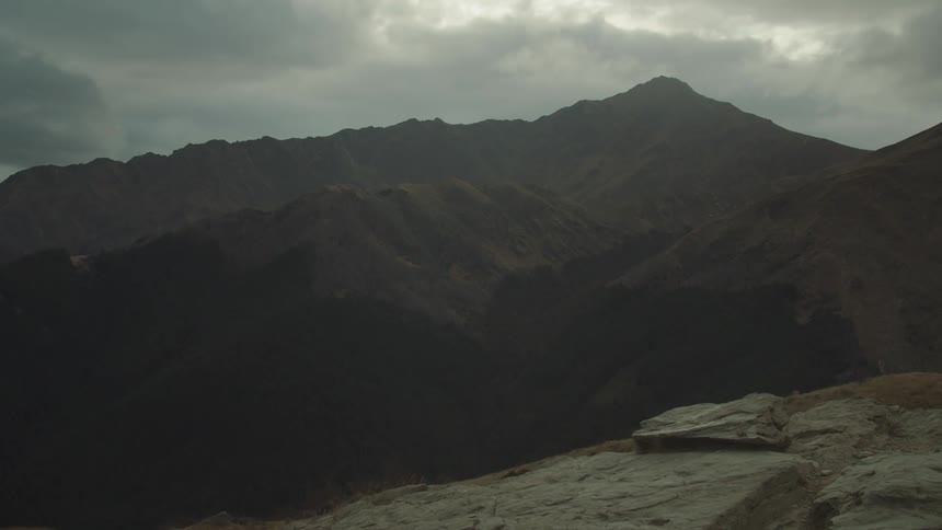 From a rocky outlook a dark rugged mountain range stretches across a valley under a heavily clouded sky