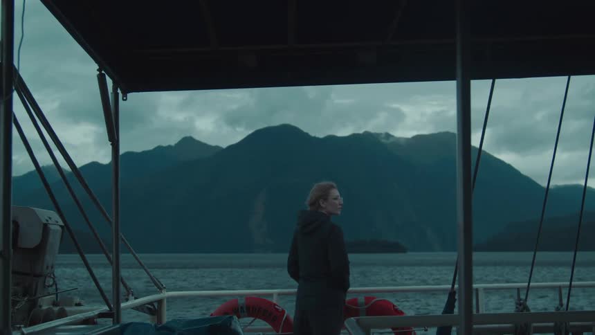 A woman standing still with her hands in her pockets on a boat gazing at the scenery the calm sea and massive mountains on the horizon
