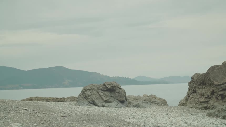 A rocky pebbled shoreline meets the calm ocean with gentle waves as distant hills are visible under an overcast sky